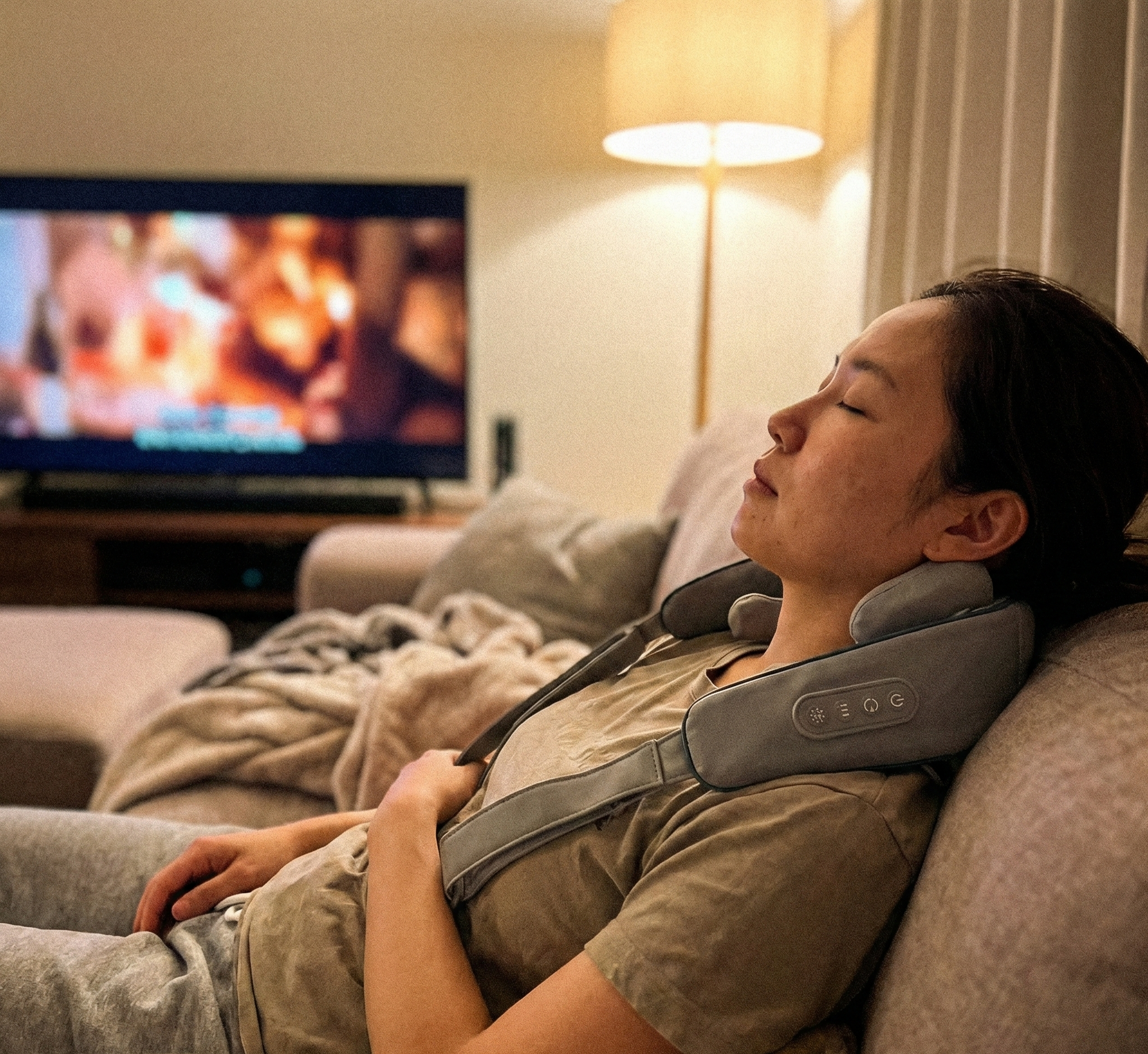 Person using a neck and shoulder massager in a living room with a TV in the background